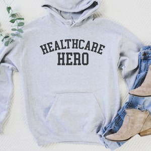 May include: A gray hooded sweatshirt with the text "Healthcare Hero" printed in black on the front. The sweatshirt is laid flat on a white surface with a pair of blue jeans and brown ankle boots.