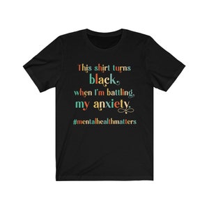 Op de afbeelding: Zwart T-shirt met de tekst "This shirt turns black when I'm battling my anxiety." en de hashtag "#mentalhealthmatters" in kleurrijke tekst.
