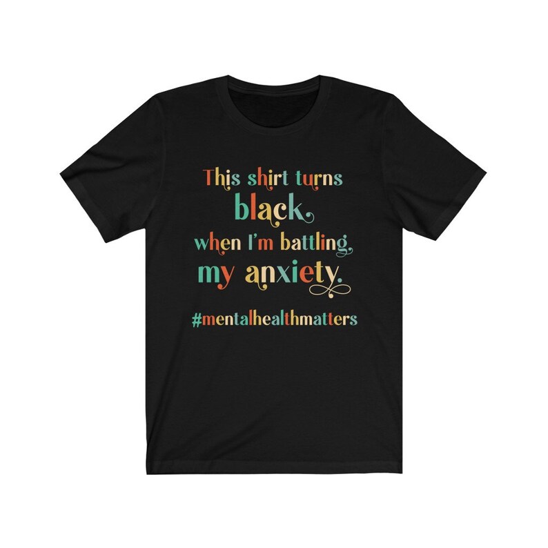 Op de afbeelding: Zwart T-shirt met de tekst "This shirt turns black when I'm battling my anxiety." en de hashtag "#mentalhealthmatters" in kleurrijke tekst.