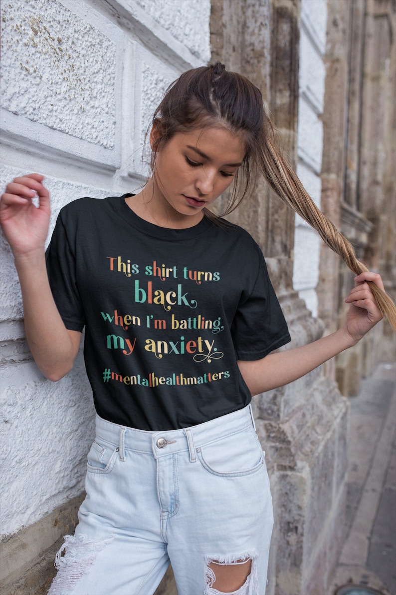 Op de afbeelding: Een zwart T-shirt met de tekst "This shirt turns black when I'm battling my anxiety" in kleurrijke letters. De tekst wordt gevolgd door de hashtag "#mentalhealthmatters".