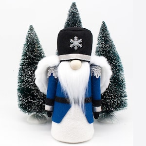 May include: A blue and white Christmas gnome dressed as a nutcracker with a black hat and silver snowflake. The gnome is standing in front of two small green Christmas trees.