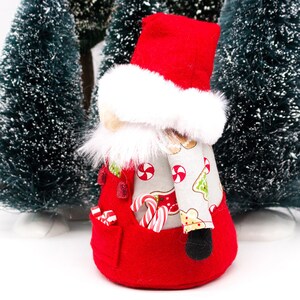 May include: A red and white Christmas gnome with a white fur trim hat and a candy cane pattern on the body. The gnome is holding candy canes in its hands.
