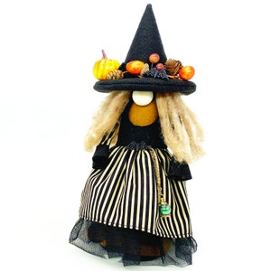 Puede incluir: Una muñeca gnomo bruja negra con un vestido a rayas blancas y negras, un sombrero de bruja negro con decoraciones de otoño y una cadena dorada con un colgante verde.