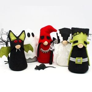 May include: Five Halloween gnomes, each dressed as a different character: a bat, a ghost, a devil, a bride, and Frankenstein. The gnomes are made of felt and have a variety of colors and patterns. A small black toy rat is in the foreground.