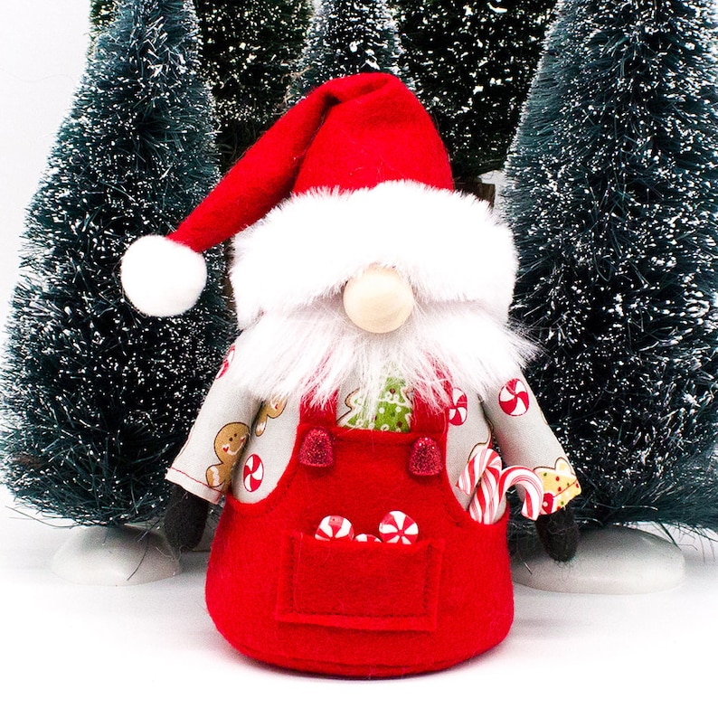 May include: A red and white Christmas gnome wearing a red hat, white beard, and a red overall with candy cane and gingerbread print. The gnome is standing in front of two artificial Christmas trees.