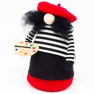 May include: A felt gnome wearing a red beret, a black and white striped shirt, and a black skirt. The gnome is holding a wooden paint palette with colourful paint.