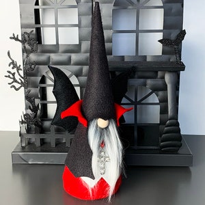 May include: A black and red Halloween gnome with a pointed hat, bat wings, and a white beard. The gnome is wearing a red and black costume with a cross pendant.