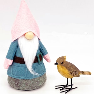 May include: A blue and pink gnome with a white beard and a brown belt stands next to a small wooden bird with brown and yellow paint.