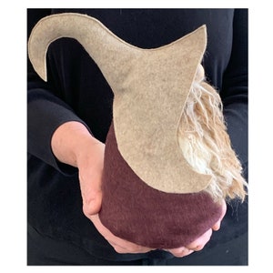 May include: A brown and beige felt gnome with a long, pointed hat. The gnome has a long, white beard and is holding a small, brown object in its hands.