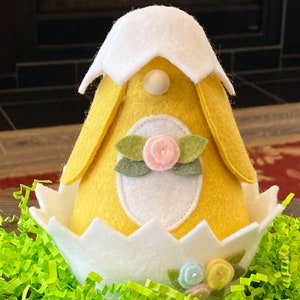 May include: A yellow felt gnome wearing a white felt hat with a white felt egg base. The gnome has a pink felt flower and green felt leaves on its chest. The gnome is sitting in a bed of green grass.
