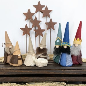May include: A felt nativity scene with a manger, baby Jesus, Mary, Joseph, and three wise men. The wise men are wearing blue, green, and red robes with gold crowns. The scene is set against a backdrop of rusty metal stars.