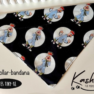 May include: Black over-collar bandana with a white polka dot pattern featuring a repeated image of a red-haired character with a surprised expression. The bandana is made of fabric and has a triangular shape. The text "Over-collar-bandana" and "SIZES TINY-XL" is printed on the bandana.