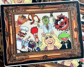 May include: A rectangular sticker featuring a framed image of various Muppet characters. The characters include Kermit the Frog, Miss Piggy, Fozzie Bear, and Animal. The frame is ornate with a gold and brown color scheme.