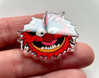May include: A clear acrylic pin featuring a vibrant red, shaggy-haired character with large white eyes, a round red nose, and a wide mouth displaying sharp white teeth. The design has a glossy finish and follows the irregular shape of the character.