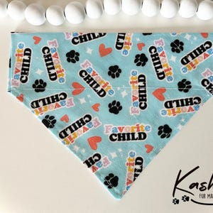 May include: A light blue dog bandana with a white background and a repeating pattern of paw prints and the text "Favorite Child" in rainbow colors.