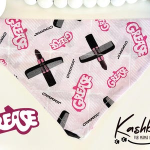 May include: Pink and white dog bandana with a repeating pattern of the word "Grease" in pink lettering, hairbrushes, lipstick, and bobby pins. The bandana is in the shape of a triangle. The word "Grease" is also printed in white letters on a pink background in the lower left corner of the image.