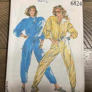 May include: A New Look 6824 sewing pattern envelope featuring illustrations of two jumpsuits. One is a solid blue long-sleeved jumpsuit with a drawstring waist and pockets. The other is a yellow and white striped long-sleeved jumpsuit with a drawstring waist.