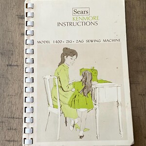 Peut inclure: Un manuel d'instructions Sears Kenmore à reliure spirale pour la machine à coudre Zig Zag modèle 1400. La couverture présente un fond blanc avec du texte noir et une illustration dans les tons verts de deux personnes à une table.