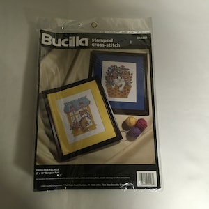 May include: A Bucilla stamped cross-stitch kit featuring two 8x10 inch samplers of cats. The kit includes pre-stamped fabric, cotton embroidery floss, needles, and easy-to-follow instructions. The kit is packaged in a clear plastic bag with a white background and the Bucilla logo.