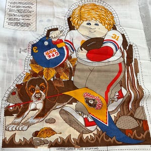 Op de afbeelding: Een wit stoffen paneel met een bedrukte Cabbage Patch Kids-figuur in een football-uniform met helm, een football en een hond die een wimpel vasthoudt. De stof bevat gestippelde kniplijnen en gedrukte naai-instructies voor het maken van een pop.