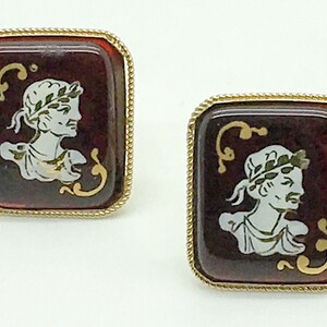 Op de afbeelding: Een paar gouden manchetknopen met een vierkante rode cameo met een wit profiel van een persoon met een lauwerkrans. De cameo is omgeven door een gouden touwachtige rand.