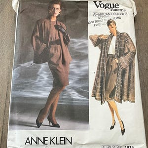 May include: A Vogue Patterns sewing pattern envelope for Anne Klein design 1915. The front displays two illustrations of outfits: a brown jacket and skirt set, and a long plaid coat over a skirt. Text includes Vogue Patterns, American Designer, and 1915.