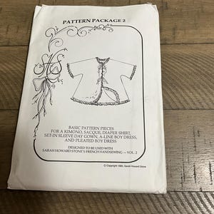 May include: A white paper sewing pattern envelope titled PATTERN PACKAGE 2. The front features a black line drawing of a ruffled garment, decorative flourishes, and text listing patterns for a kimono, sacque, diaper shirt, day gown, and dresses.
