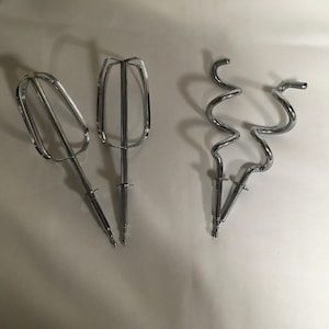 May include: Three shiny, chrome-colored mixer attachments are displayed against a white background. The attachments include a whisk, a beater, and a dough hook, all designed for use with a hand mixer. These are essential baking tools.