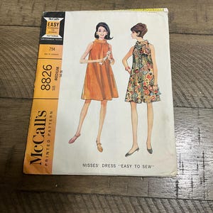 May include: Vintage McCall's printed sewing pattern for Misses' dresses. The pattern features two dress designs, one in orange and the other in a floral print. The pattern is labelled "Easy to Sew" and includes the number 8826.