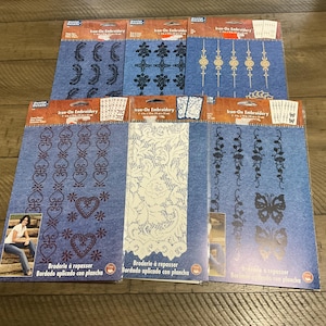 May include: A collection of iron-on embroidery patches in various designs, including hearts, floral patterns, and butterflies. The patches are on blue fabric, with some featuring a woman in jeans. Each package is approximately 7 inches by 10 inches.