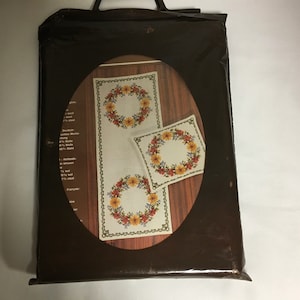 May include: A set of three white embroidered table linens featuring a floral wreath design in shades of yellow, orange, and red. The linens are packaged in a brown plastic bag.