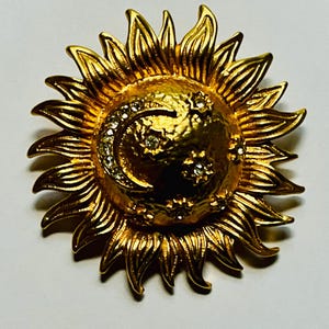 May include: A gold-toned metal brooch shaped like a sun with radiating pointed rays. The center features a textured surface with a crescent moon and several small stars, each inlaid with clear, sparkling rhinestones that catch the light.