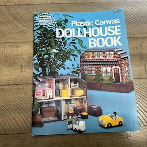 Op de afbeelding: Een instructieboek met zachte kaft getiteld Plastic Canvas DOLLHOUSE BOOK 3008. De lichtblauwe kaft toont een foto van een bakstenen poppenhuis met meerdere verdiepingen, ingerichte kamers, een gele speelgoedauto en twee kleine figuren van plastic canvas.