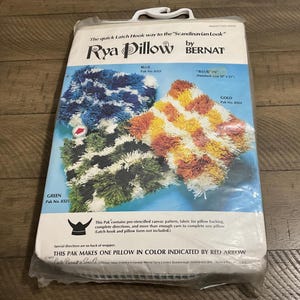 May include: A Bernat Rya Pillow latch hook kit in a plastic package. The packaging displays three finished pillow designs in blue, green, and gold colorways, with text indicating a finished size of 12 inches by 15 inches and a red arrow.