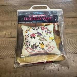 Op de afbeelding: Een vintage Avon Creative Needlecraft crewel borduurpakket. De set bevat een kussen met een bloemenontwerp dat de Dertien Originele Koloniën vertegenwoordigt. Het kussen is mesvormig en vierkant. De set is verpakt in een doorzichtige plastic zak.