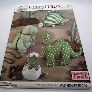 May include: A Simplicity Crafts sewing pattern for making stuffed dinosaurs. The pattern includes instructions for making a green and white striped dinosaur, a green and white polka dot dinosaur, and a baby dinosaur in an egg. The pattern is from 1987 and is by Shirley Botsford.
