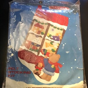 May include: A felt stocking kit in a clear plastic package. The stocking design features a teddy bear, houses, and other festive elements. The stocking measures 15 inches in length. The text "FELT STOCKING KIT" is visible.
