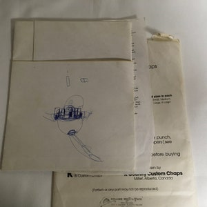 Op de afbeelding: Een witte envelop met een handgetekend cartoonfiguur erop. De envelop heeft tekst die "A & Custom Chaps" en "Custom Chaps" luidt met het adres "Millet, Alberta, Canada". De tekst zegt ook "4 maten in elke" en somt de maten op als "Small, Medium, Large, X-Large".