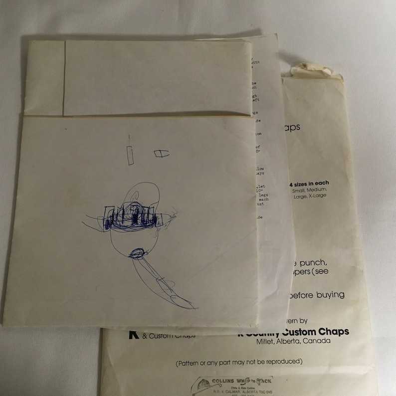 Op de afbeelding: Een witte envelop met een handgetekend cartoonfiguur erop. De envelop heeft tekst die "A & Custom Chaps" en "Custom Chaps" luidt met het adres "Millet, Alberta, Canada". De tekst zegt ook "4 maten in elke" en somt de maten op als "Small, Medium, Large, X-Large".