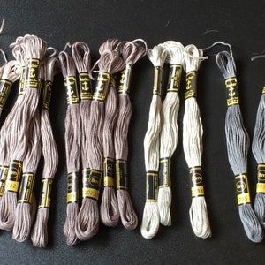 May include: A collection of Anchor embroidery floss skeins in neutral shades of beige, taupe, gray, and olive green. Each skein features a black and yellow paper label with the brand name Anchor and an anchor logo, along with specific color numbers.