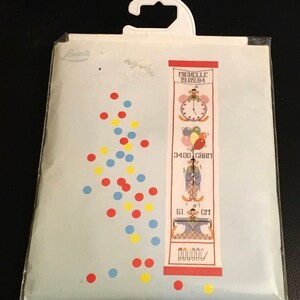 May include: A blue and white cross-stitch pattern with a clown theme. The pattern includes a clock, balloons, and a clown holding a measuring tape. The text on the pattern reads "Michelle 19.09.84", "3400 GRAM", "51 CM", and "DOUDOU".