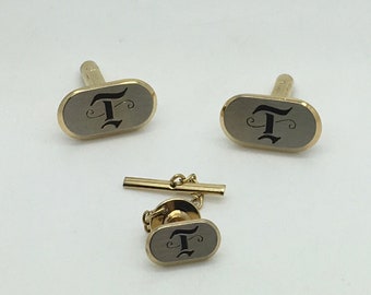 May include: A set of three gold-toned cufflinks and a tie tack. Each piece features a black enamel oval with a stylized letter 'T' in the center.