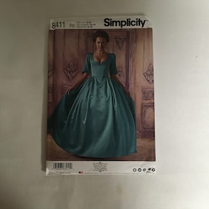 Könnte beinhalten: Simplicity Schnittmuster Nummer 8411, Größe US 14-22, Talla Euro 40-48, Taille Fr 42-50, für ein türkisfarbenes Kleid mit tiefem Ausschnitt und einem taillierten Oberteil. Das Model trägt das Kleid vor einer dekorativen Wand.