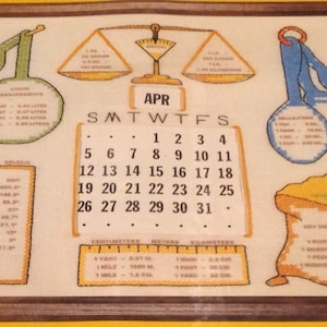 May include: Cross-stitch kitchen-themed artwork featuring measurement conversions, a calendar for April, and a thermometer. Includes illustrations of measuring spoons, scales, and a bag of flour. The artwork is framed in a wooden frame.