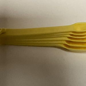Peut inclure: Un ensemble de cinq cuillères à mesurer jaunes, imbriquées les unes dans les autres. Les cuillères sont en plastique et ont une forme arrondie et lisse.