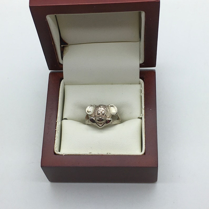 May include: A silver ring featuring a Mickey Mouse head design, presented in a brown ring box.