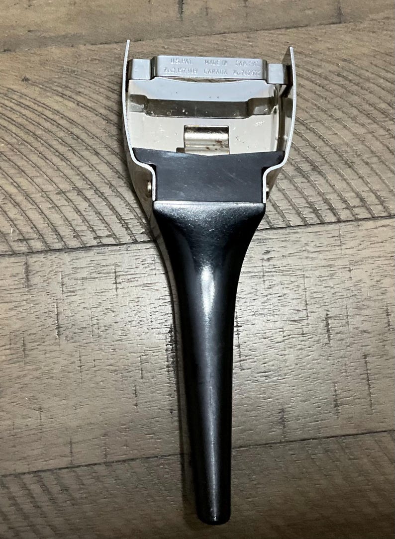 May include: A metal vegetable peeler with a black handle. The top metal section features a horizontal blade and is stamped with the text 'REGAL MADE IN CANADA' and 'PATENT CANADA NO 742072'. The tool has a tapered handle and a wide, U-shaped head.