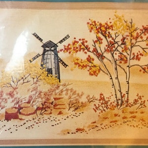 May include: Embroidered picture of a windmill with a white and black design, surrounded by trees with yellow and red leaves. The background is a light brown color.