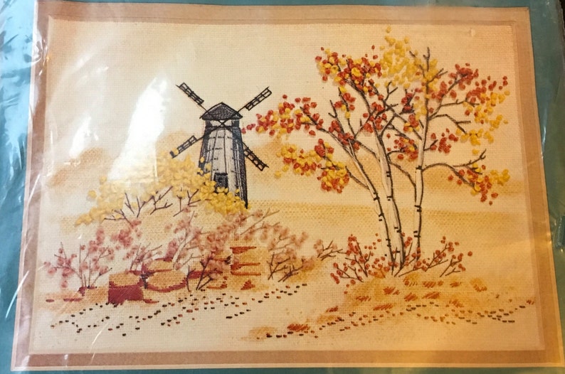 May include: Embroidered picture of a windmill with a white and black design, surrounded by trees with yellow and red leaves. The background is a light brown color.