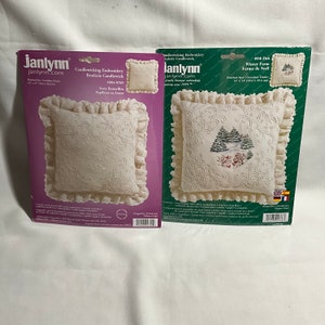 Op de afbeelding: Twee pakketten met borduursets voor kussens. Het linker pakket bevat een wit kussen met een ivoor kleurig vlindermotief en de tekst "Candlewicking Embroidery Broderie Candlewick 2006-0769 Ivory Butterflies Papillons en Ivoire". Het rechter pakket bevat een wit kussen met een winterscene van bomen en de tekst "Winter Farm Ferme de Noel 104-766 Finished Size/Grandeur Finie 14" x 14"/35.6cm x 35.6cm".